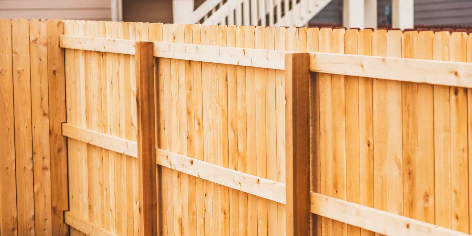 Wooden fence with vertical planks and horizontal supports, painted in light, natural wood tones. Blurred background of a residential building with a staircase. Crafted expertly by Fencing Services Duval County FL for a blend of style and durability.