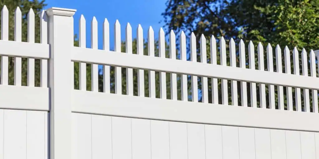 A white vinyl fence with pointed pickets stands proudly under a clear blue sky. Featuring a clean, modern design, it contrasts beautifully with lush green trees in the background. Discover this craftsmanship through Fencing Services Duval County FL.