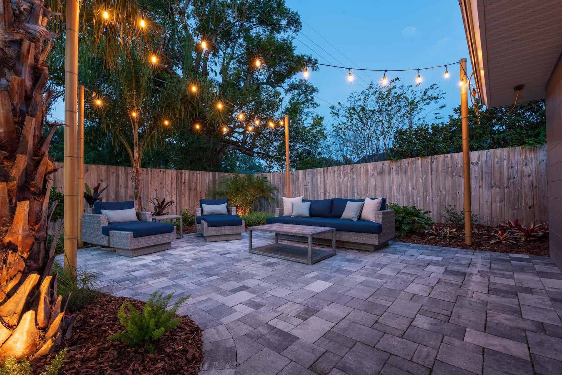 A cozy outdoor patio at dusk features wicker sofas with blue cushions and white pillows. String lights hang above, creating a warm ambiance. The area is highlighted by expert fencing installation from a trusted Duval County, FL company, surrounded by lush greenery, with a paver stone floor.