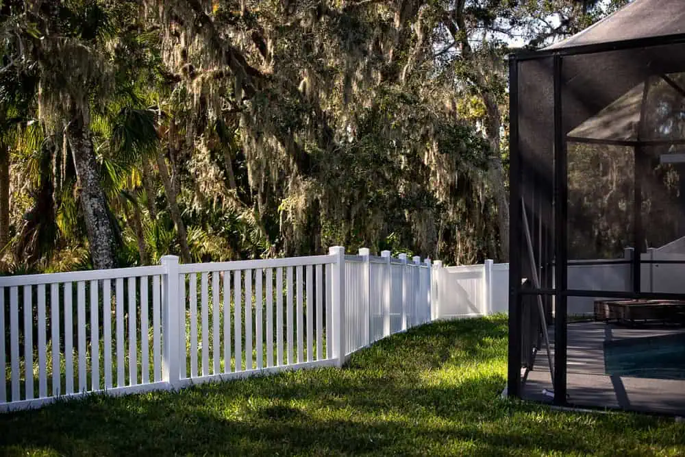 A white picket fence by a reputable fencing company in Duval County, FL, borders a lush yard with trees draped in moss. On the right, a screened patio is partially visible, casting shadows on the grass. The sunlit scene creates a serene outdoor setting.