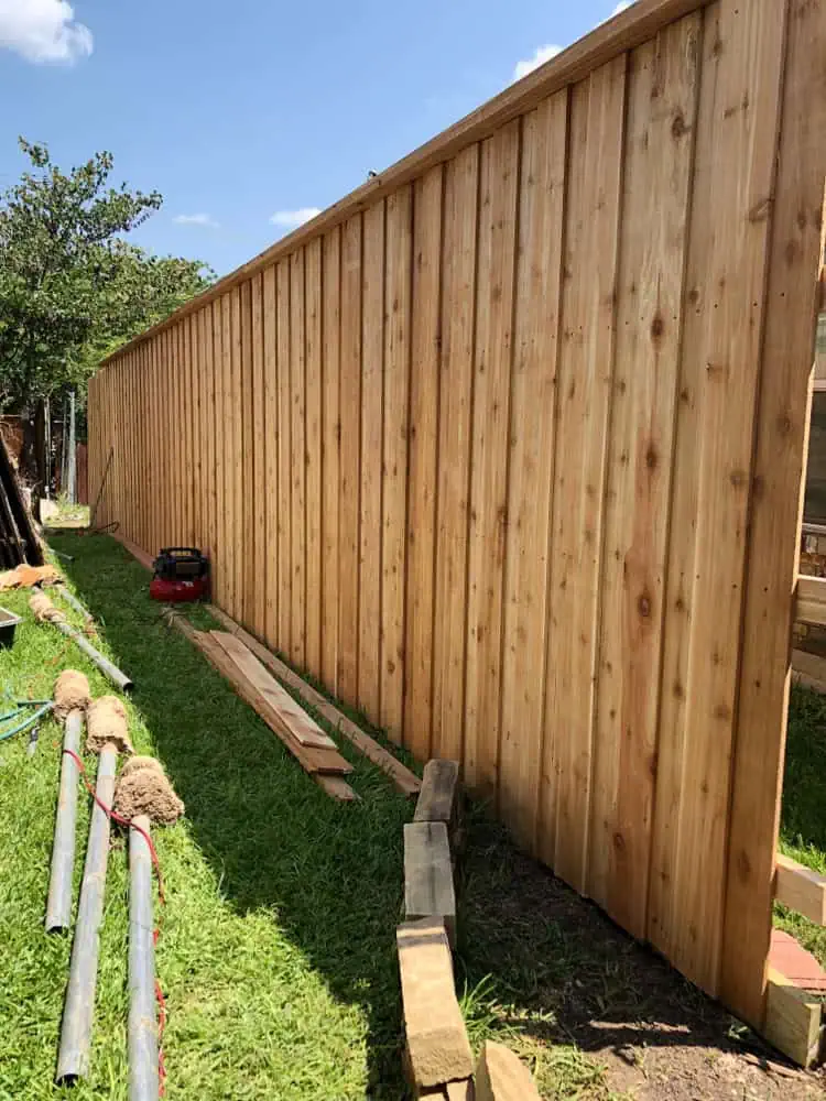A long wooden fence is being constructed on a grassy area under a clear blue sky in Duval County, FL. Building materials, such as pipes and wooden planks, are scattered nearby. A red toolbox and some construction tools are on the ground, showcasing expert fencing installation services.