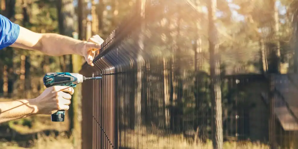 A person uses a cordless drill to secure a metal fence under the bright sun, with trees blurred in the background. Ideal for those seeking quality fencing installation, Duval County FL offers top-notch expertise for your outdoor projects.