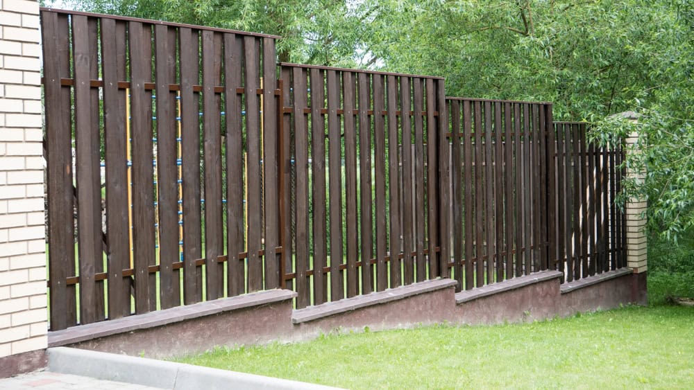 Wooden fence with vertical slats and brick pillars, set on a sloped concrete base by Fencing Installation Duval County FL. The fence is surrounded by greenery, with trees in the background and grass in the foreground.