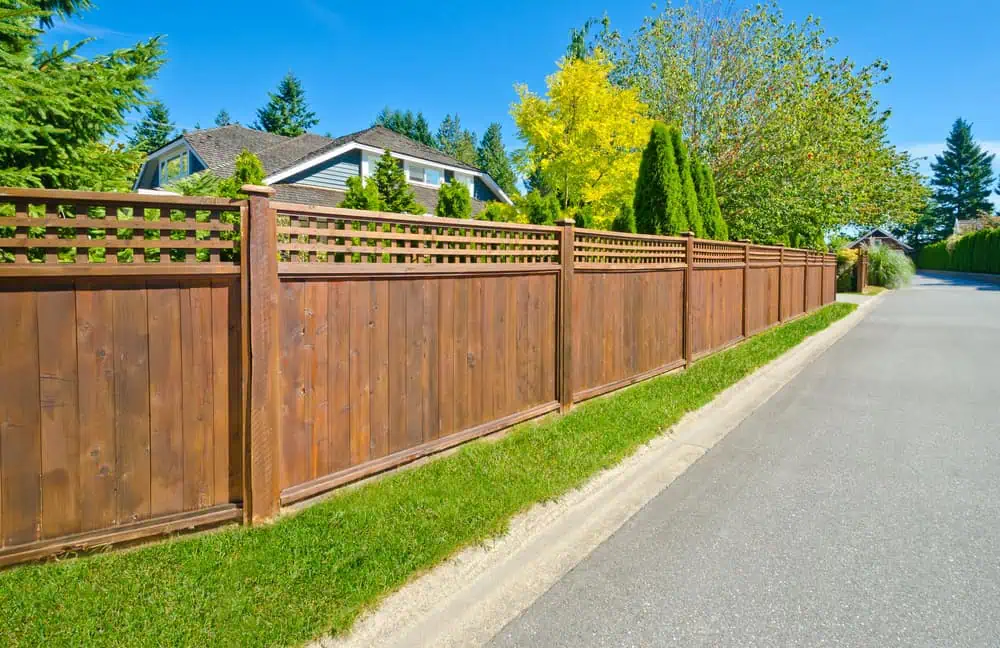 A long wooden fence lines a sidewalk, with green grass on one side. Behind the fence, there's a house partially visible with trees and blue sky in the background—showcasing the craftsmanship of a top-notch fencing company in Duval County, FL.