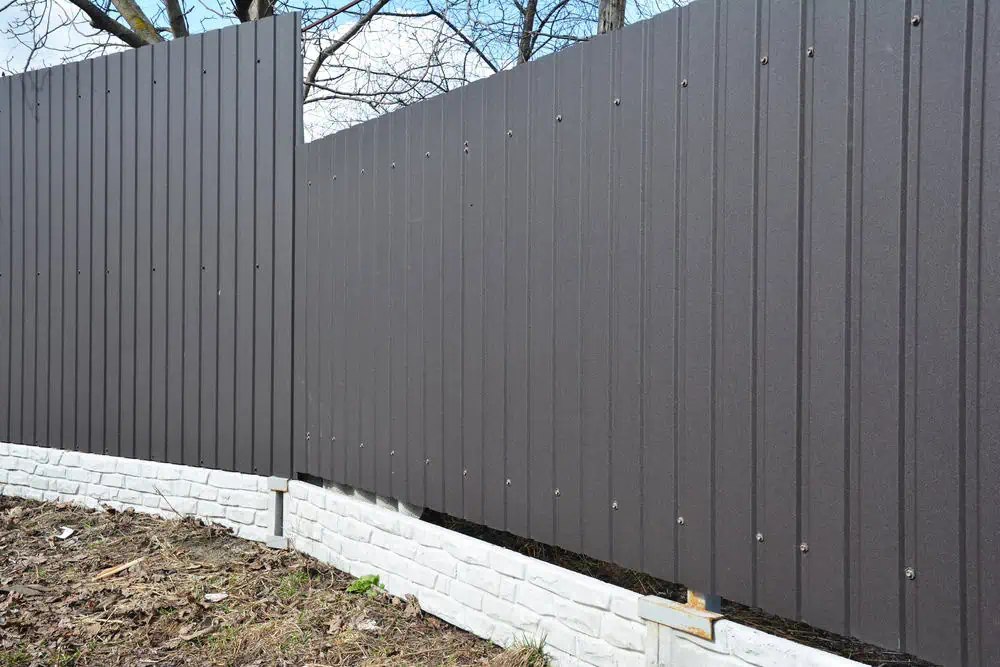 A modern gray metal fence, expertly installed by a fencing company in Duval County, FL, stands atop a short white brick wall. The fence panels align neatly with a small section creating an angle. In the background, tree branches are visible against a cloudy sky, with dry leaves covering the ground.