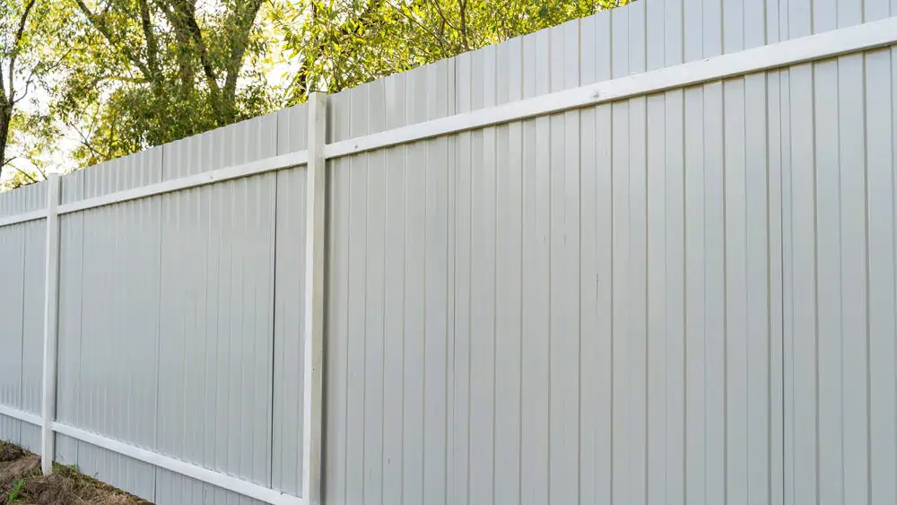 Tall white vinyl fence with vertical slats and posts, expertly installed by a trusted fencing company in Duval County, FL. Set against a backdrop of green trees with sunlight filtering through leaves, it offers both beauty and privacy to your outdoor space.