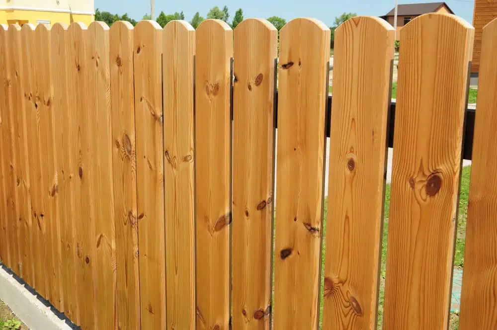A wooden picket fence with vertical planks showcases a natural finish and visible wood grain. Set outdoors amidst green grass under a blue sky, this elegant design is expertly crafted by Fencing Services Duval County FL.