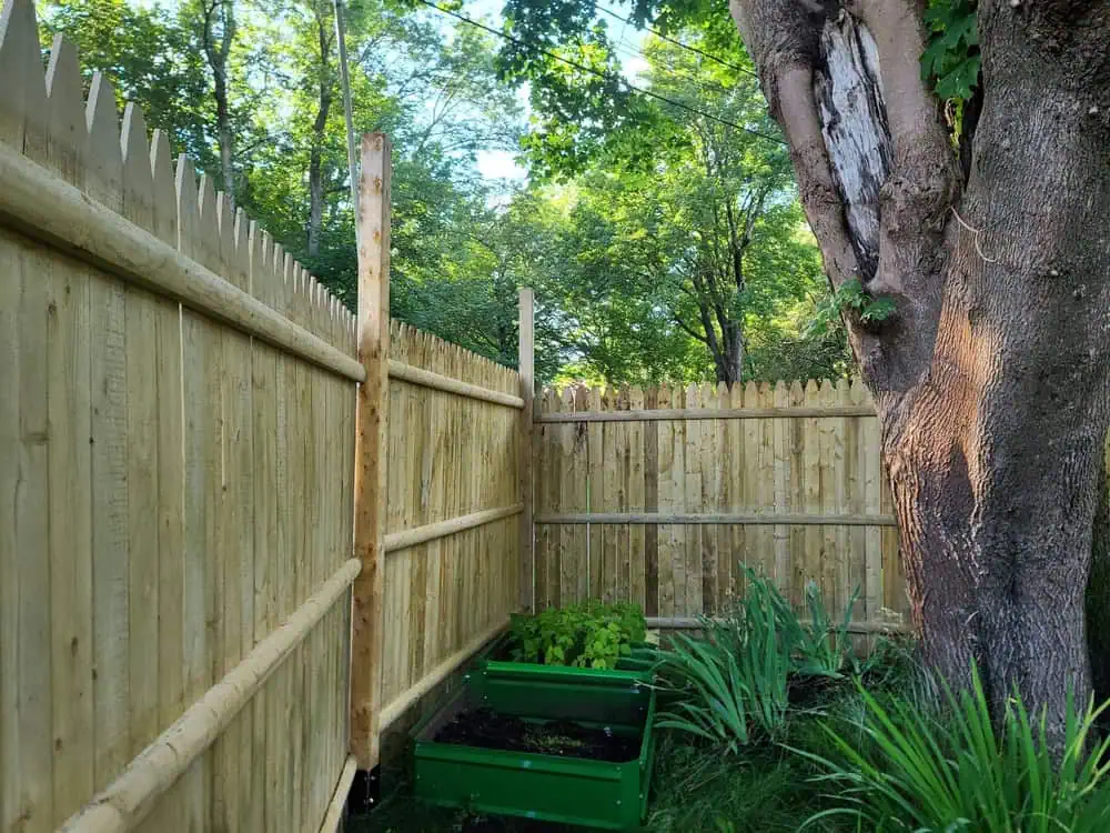 A wooden fence corner, expertly crafted by a fencing company in Duval County, FL, surrounds a garden with two green planter boxes. The fenced area boasts lush foliage, and a large tree grows nearby, partially visible on the right. The scene is bright and sunny.