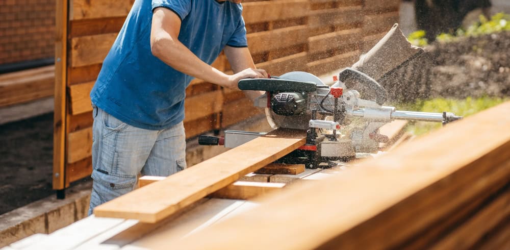 A person in a blue shirt expertly uses a circular saw to cut a wooden board outdoors, surrounded by sawdust and planks. A wooden fence hints at their craftsmanship, likely for Fencing Installation in Duval County, FL.