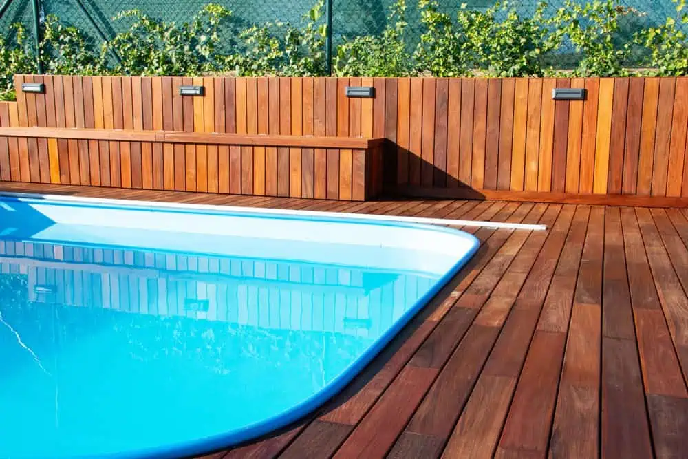 A modern outdoor space features a swimming pool with light blue water and a surrounding deck of rich, dark wooden planks. A sleek wooden wall with built-in bench seating lines one side, adorned with greenery, seamlessly complemented by expert fencing services in Duval County, FL.