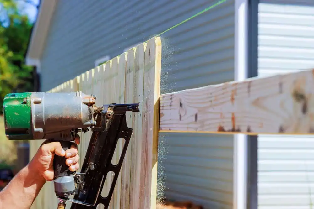 A person uses a pneumatic nail gun to secure vertical wooden planks to a horizontal beam, constructing a fence with precision as expected from Fencing Installation Duval County FL. A green guiding line ensures alignment, and in the background, a white siding building is visible.