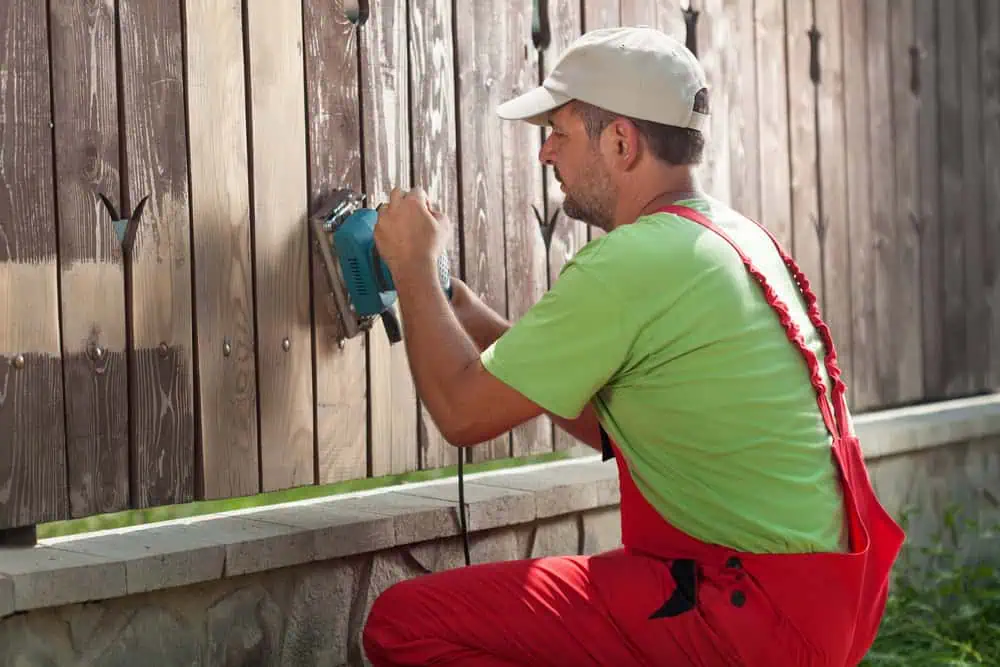 A man in a green shirt and red overalls, from a Fencing Company in Duval County FL, uses an electric sander on a wooden fence. Wearing a beige cap, he is working outdoors with grass visible in the background. The fence sits atop a stone base.