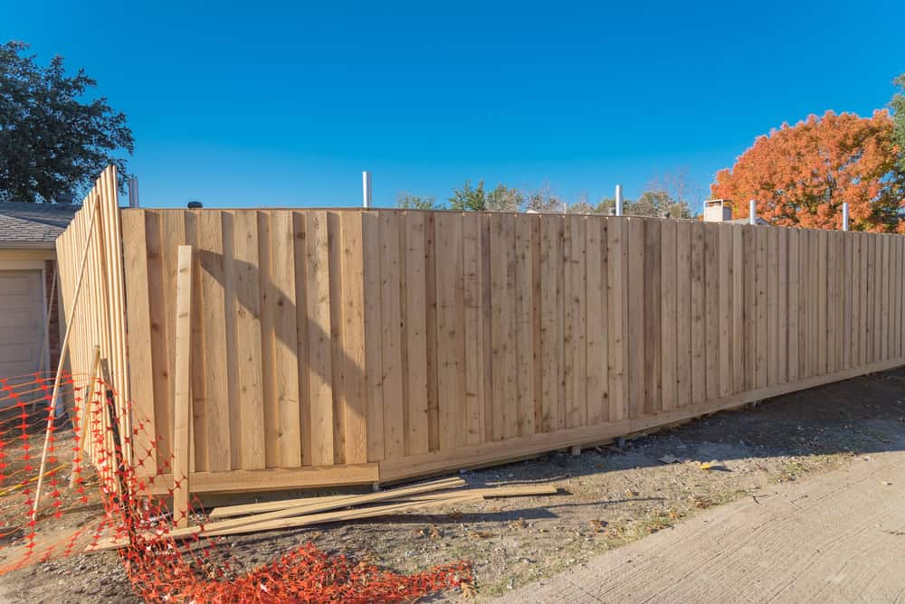 A tall wooden fence under construction, with vertical planks, stands beside an outdoor area. Orange safety netting is visible in the foreground, and trees with autumn foliage are in the background under a clear blue sky—a testament to expert fencing installation in Duval County, FL.