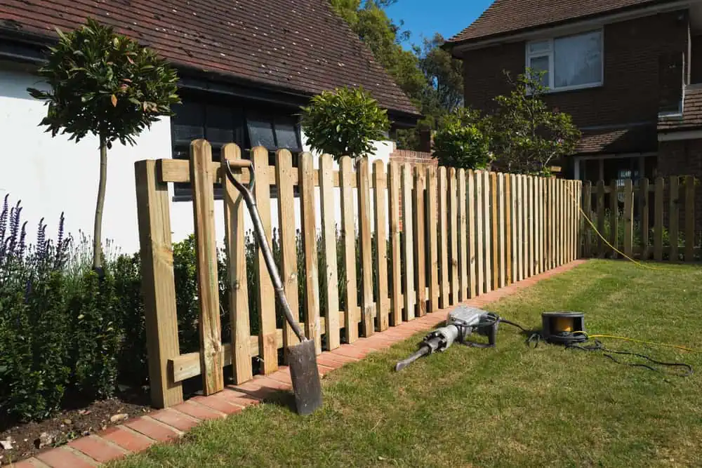 A newly installed wooden fence lines a grassy yard beside a house, showcasing impeccable craftsmanship from a leading fencing company in Duval County, FL. A shovel, power tool, and yellow extension cord are on the grass. Small trees near the fence complement the scene, with a white building in view.