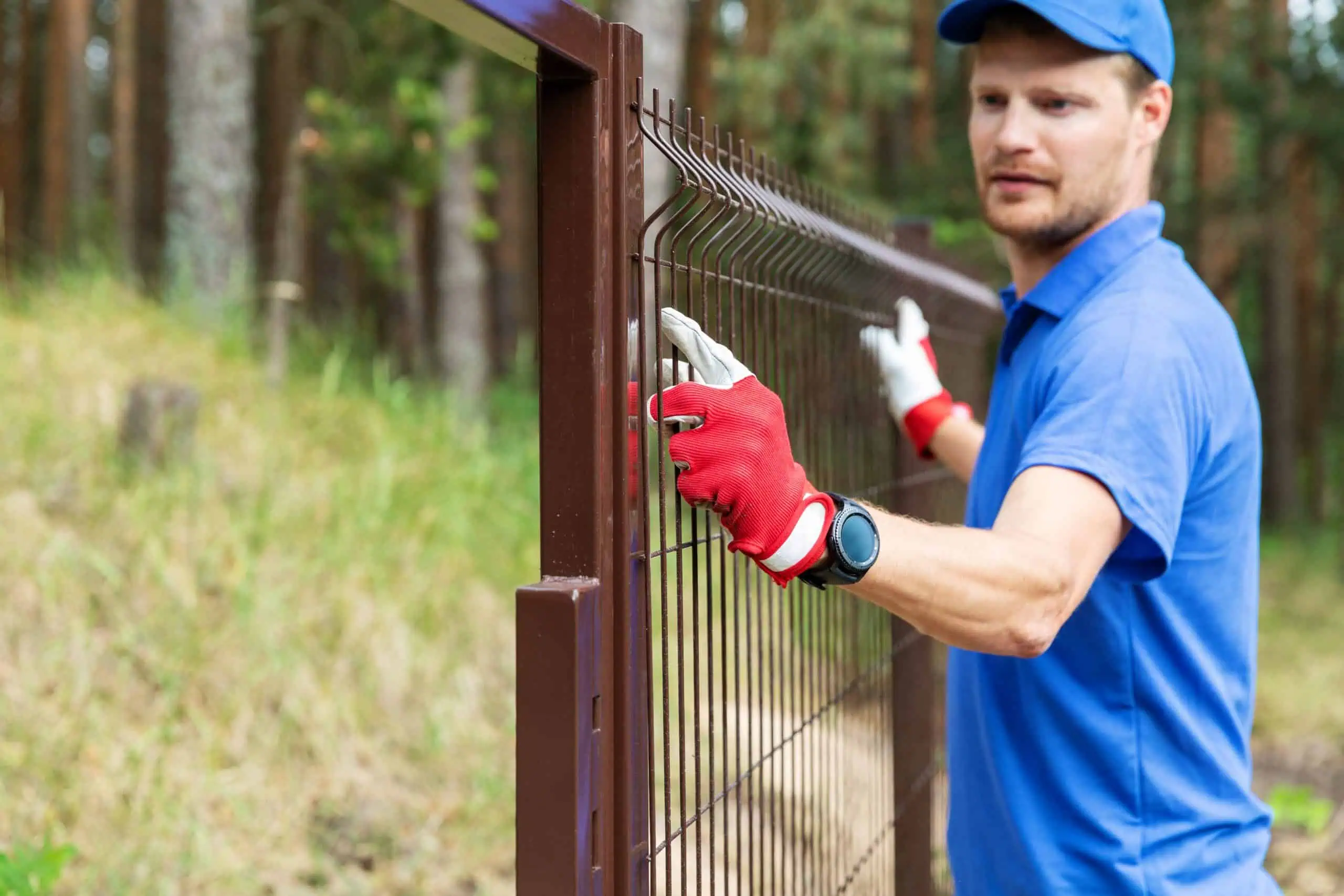 A man in a blue uniform and red gloves is expertly adjusting a metal fence amidst the lush greenery, showcasing top-notch fencing installation skills in Duval County, FL.