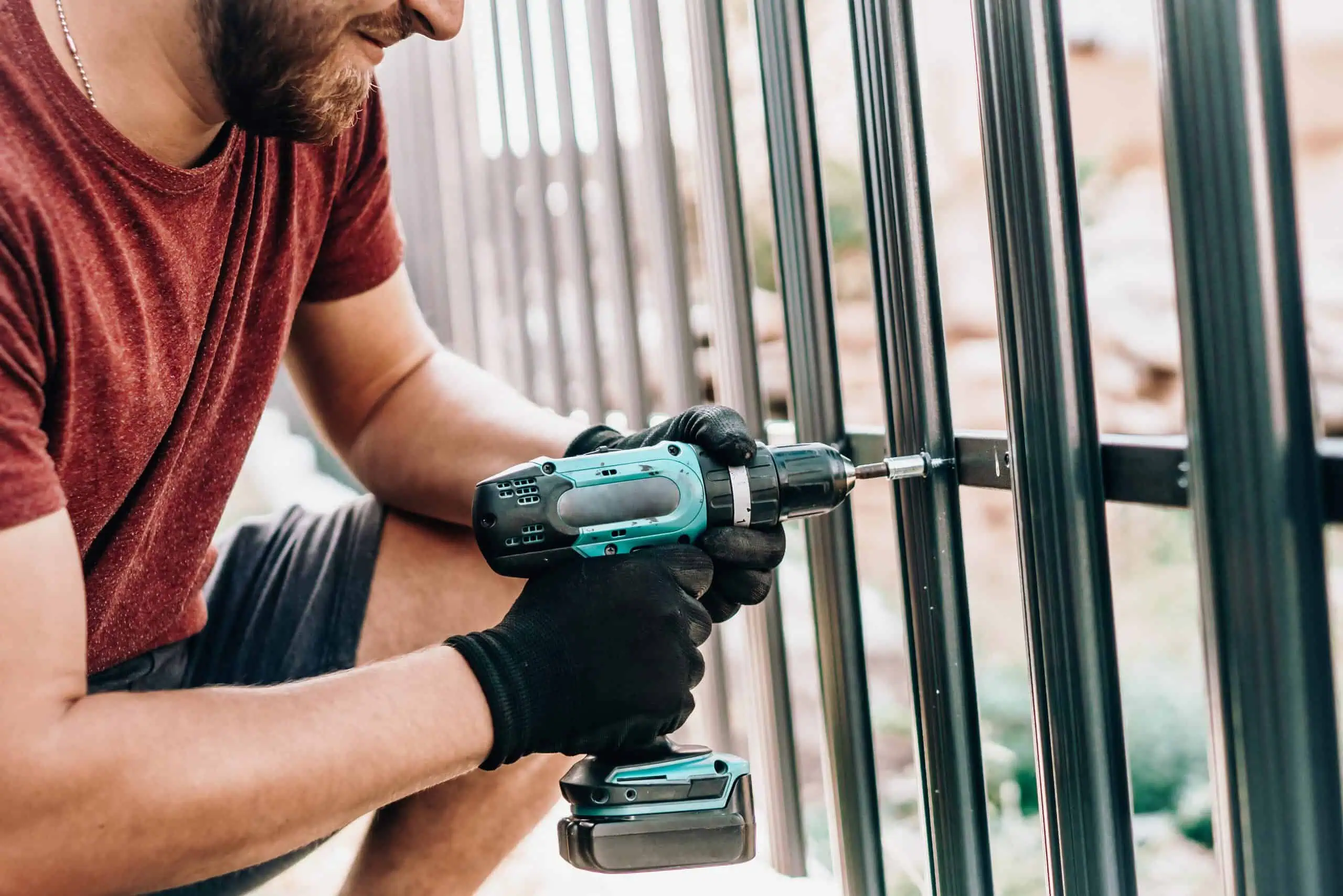 A person in a red shirt and black gloves skillfully uses a turquoise cordless drill to tighten screws on a metal fence. This fencing contractor from Duval County, FL, is working outdoors with lush greenery softly blurred in the background.