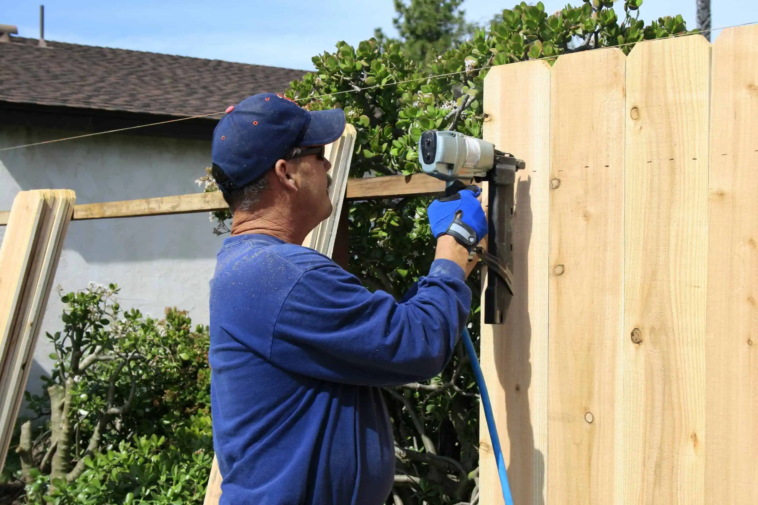 A man in a blue shirt and cap uses a nail gun to install wooden fence panels as part of professional fencing installation in Duval County, FL. Bushes are visible in the background, and the weather is clear.