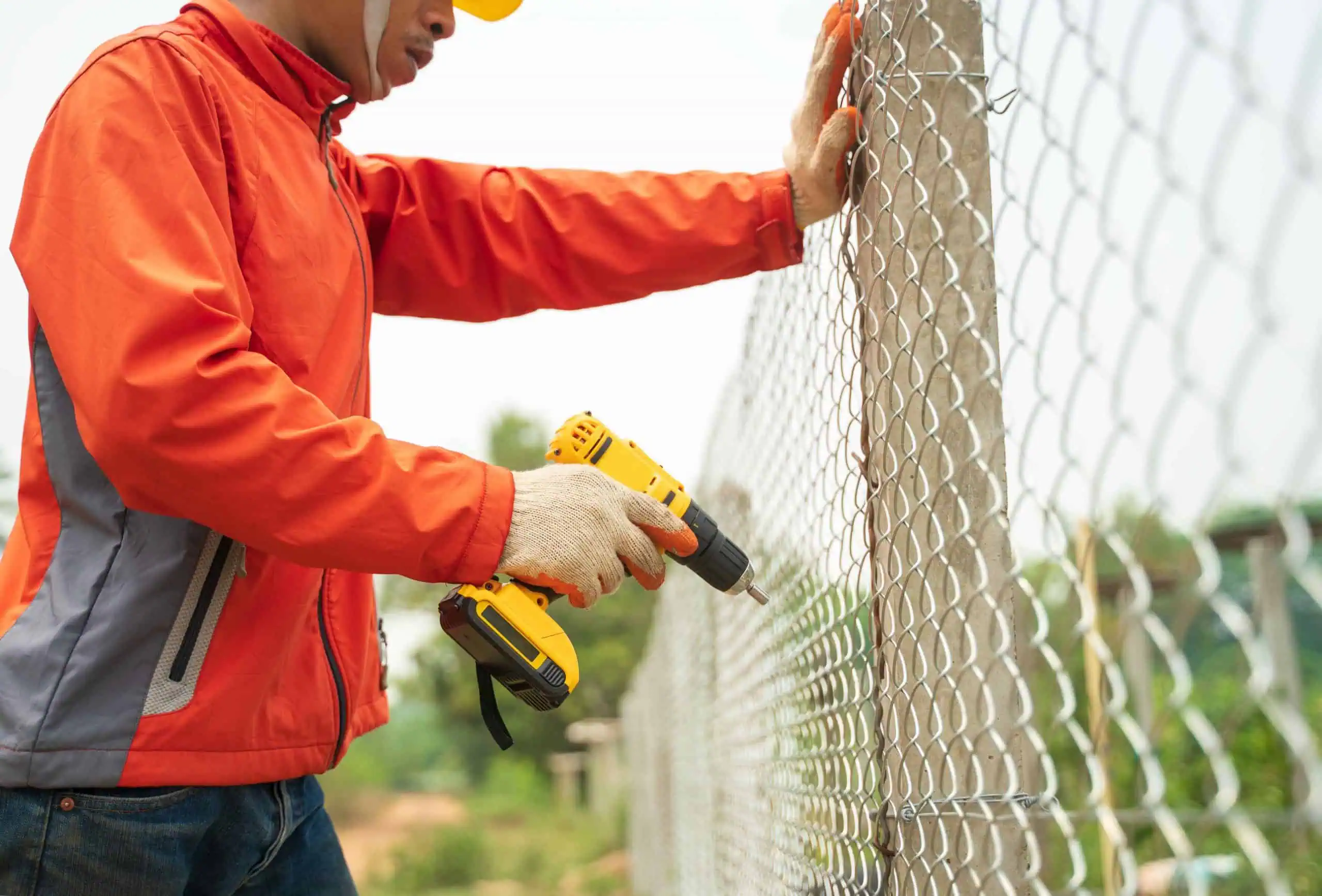 A person wearing an orange jacket and gloves uses a yellow power drill to secure a metal chain-link fence to a post, showcasing expert fencing installation in Duval County, FL. The background is blurred, highlighting lush greenery.