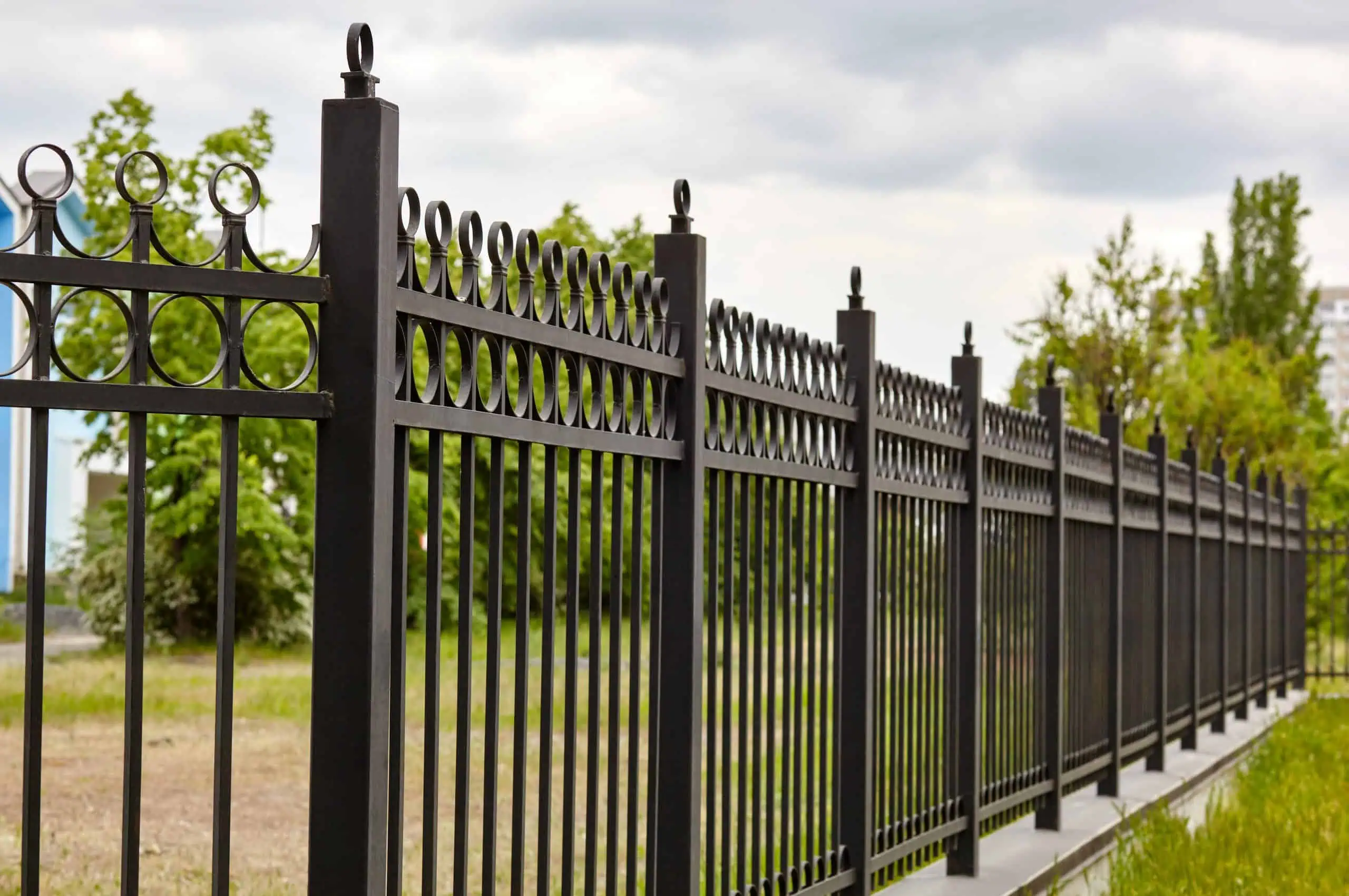 A long, decorative black metal fence, perfect for any Fencing Installation in Duval County FL, lines a grassy area with trees and a cloudy sky in the background. The fence features vertical bars and circular patterns at the top.